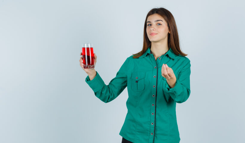 young girl holding glass of red liquid, showing Italian gesture in green blouse, black pants and looking satisfied. front view.